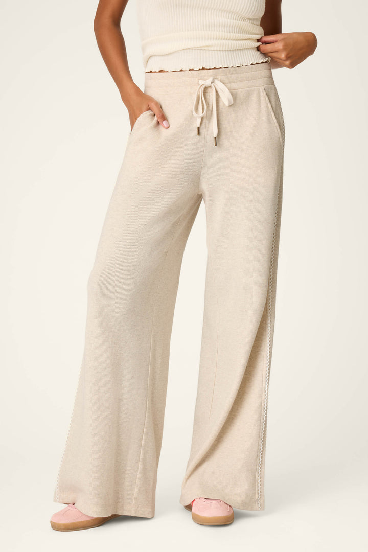A person wears the P.J. Salvage Weekday Waffles Oatmeal Lounge Pants with a drawstring waist, paired with a white ribbed crop top and light pink shoes. One hand is tucked into a pocket while the other holds the waistband. The background is light and plain.