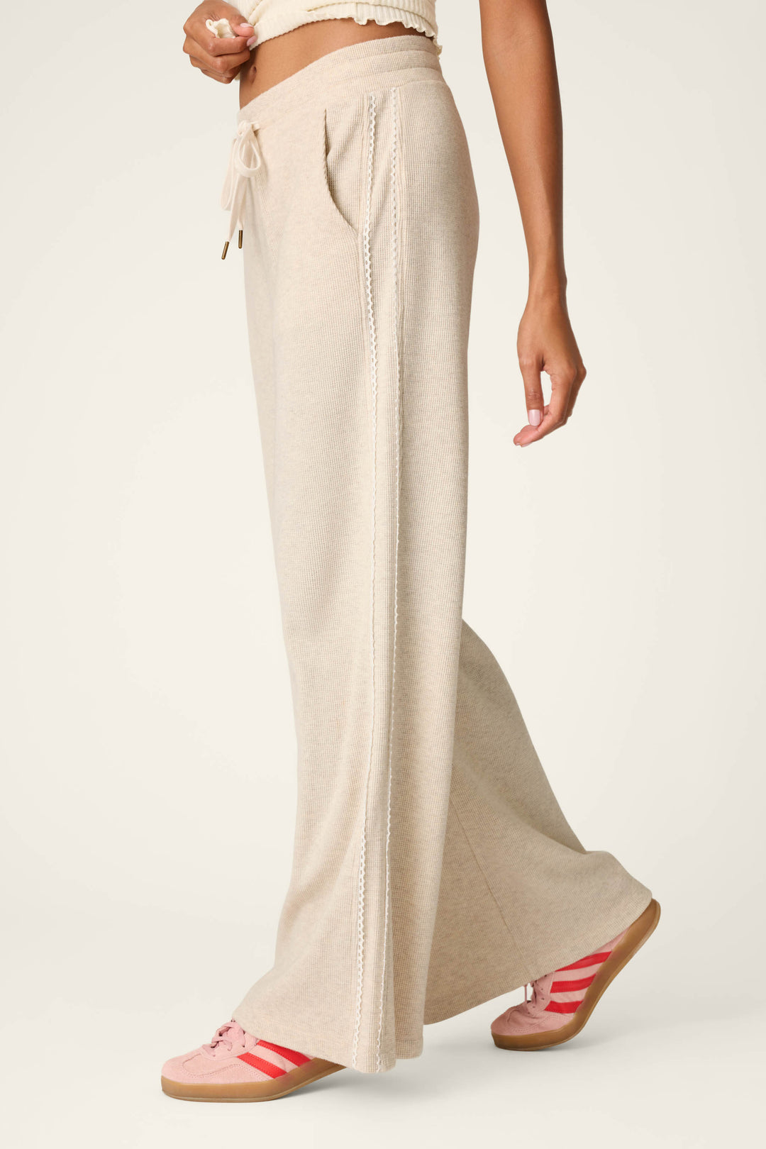A person wearing P.J. Salvage's Weekday Waffles Oatmeal Lounge Pants, paired with a cream top and pink sneakers featuring red stripes, stands against a plain light background, with only the lower body visible.