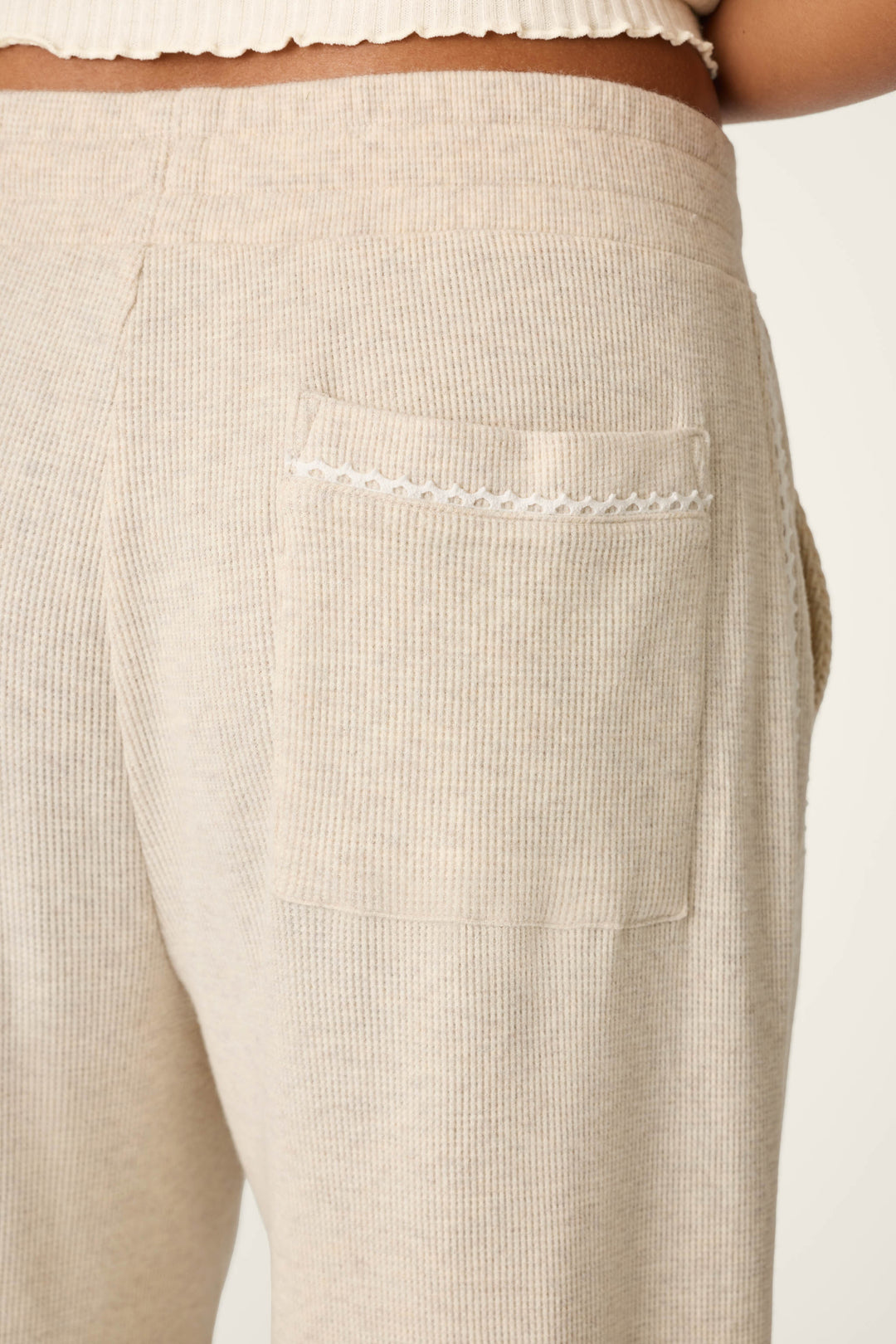 Close-up of the back pocket of the P.J. Salvage Weekday Waffles Oatmeal Lounge Pants, highlighting a small decorative scalloped trim on the pocket. The beige ribbed pants are worn by a person, with part of their waist and hip visible in the image.