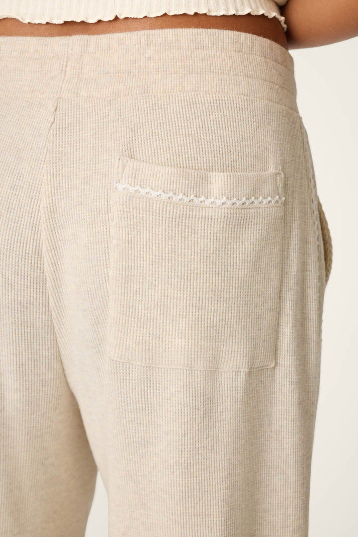 Close-up of the back pocket of the P.J. Salvage Weekday Waffles Oatmeal Lounge Pants, highlighting a small decorative scalloped trim on the pocket. The beige ribbed pants are worn by a person, with part of their waist and hip visible in the image.