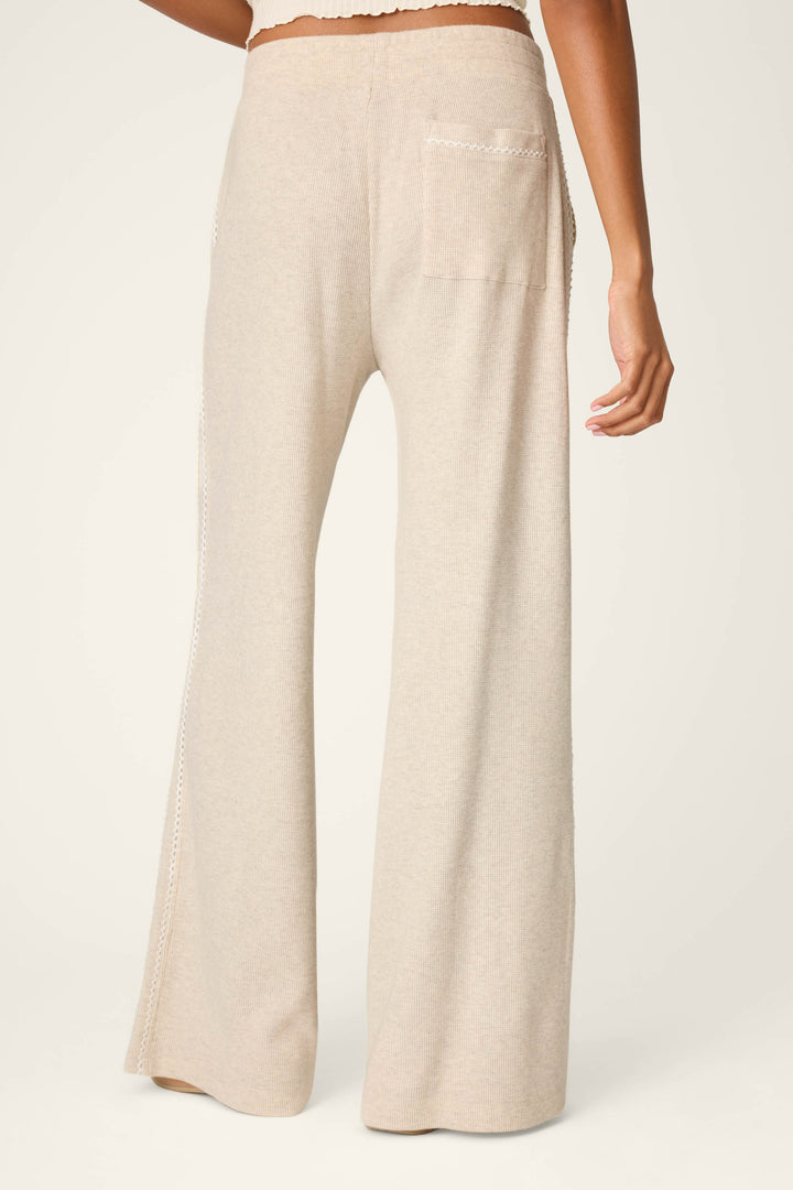 A person wearing the P.J. Salvage Weekday Waffles Oatmeal Lounge Pants stands facing away, highlighting the pants’ high-waisted fit, back pocket, scalloped trim, and relaxed wide-leg silhouette in a light beige knit fabric.