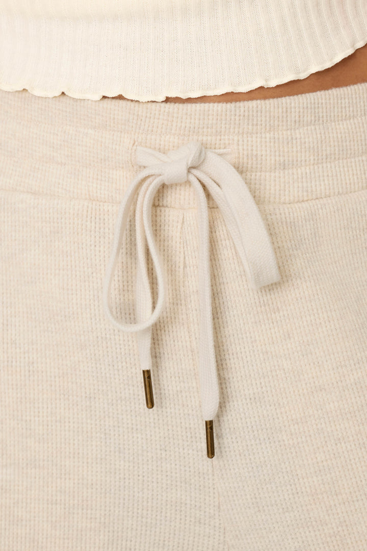 Close-up of the cream-toned ribbed waistband of the P.J. Salvage Weekday Waffles Oatmeal Lounge Pants, featuring a drawstring tied in a bow with gold-colored metal tips. The top of the pants and the hem of a ribbed white shirt are also visible.
