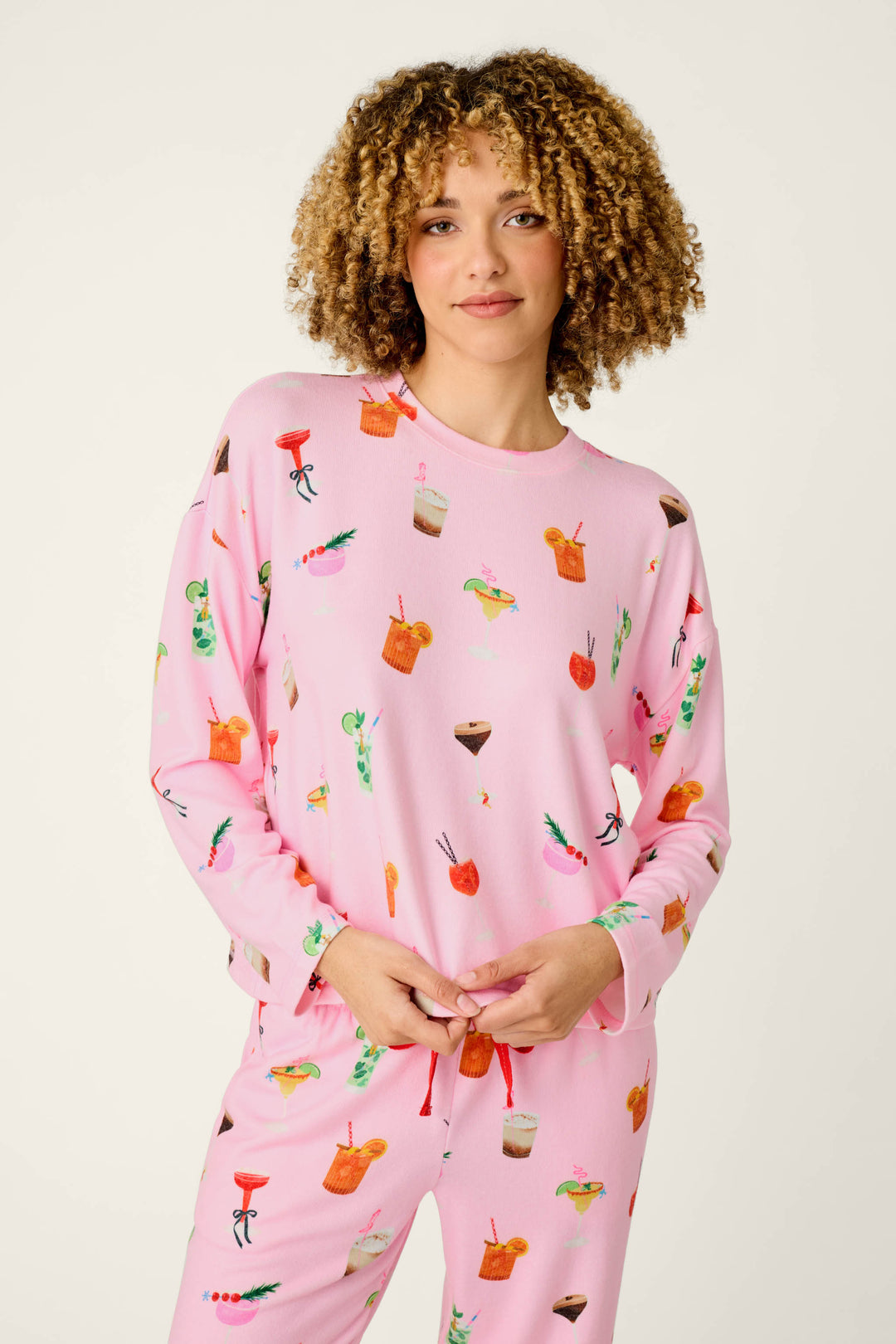 A person wearing the P.J. Salvage First Chair, Last Call Cocktail Print Long Sleeve Top, featuring colorful cocktail and drink illustrations, stands against a plain light background and smiles slightly.