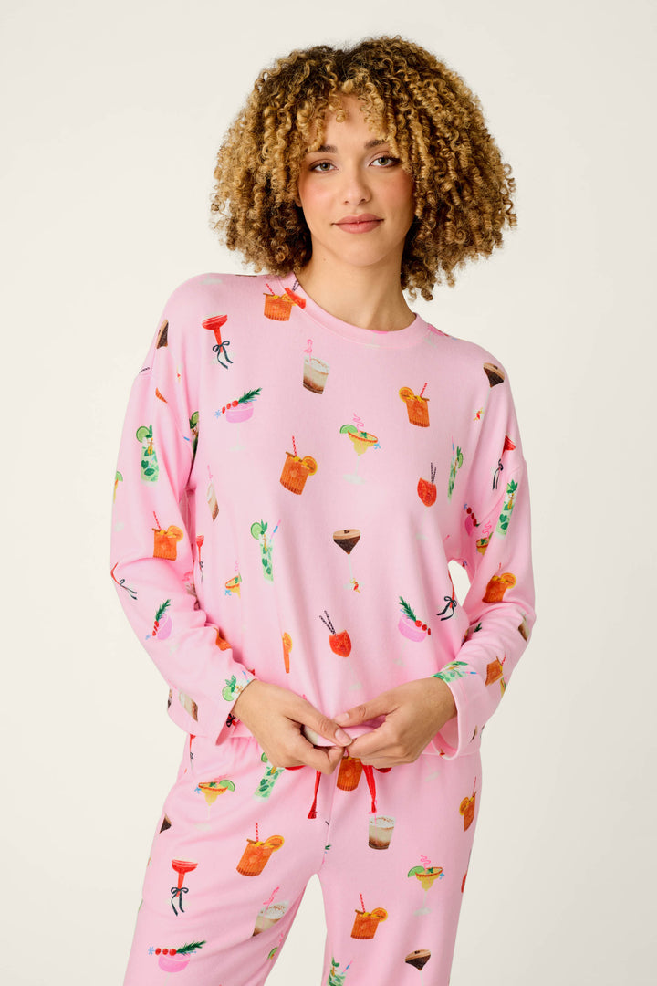 A person wearing the P.J. Salvage First Chair, Last Call Cocktail Print Long Sleeve Top, featuring colorful cocktail and drink illustrations, stands against a plain light background and smiles slightly.