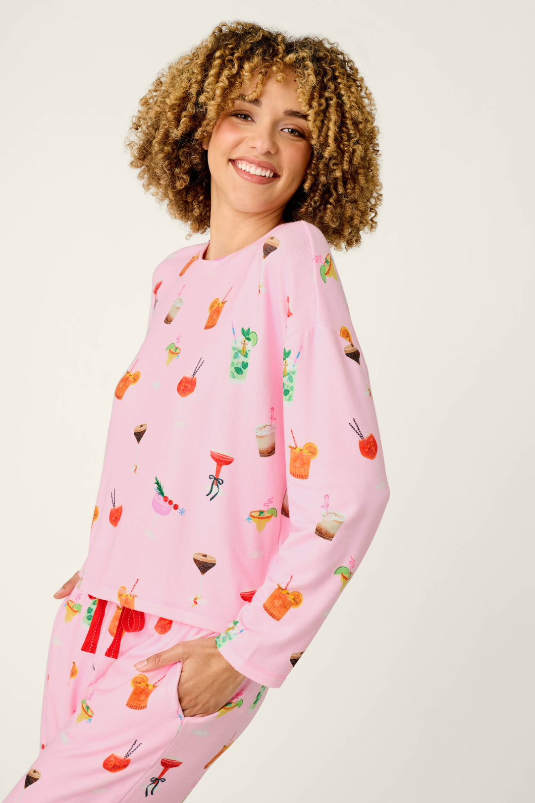 A smiling person with curly hair wears the P.J. Salvage First Chair, Last Call Cocktail Print Long Sleeve Top, featuring pink fabric decorated with colorful drink illustrations, and stands against a plain light background.