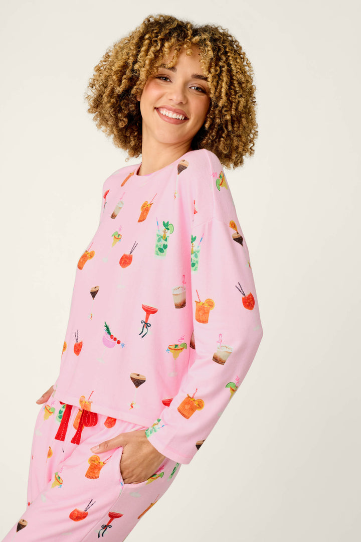 A smiling person with curly hair wears the P.J. Salvage First Chair, Last Call Cocktail Print Long Sleeve Top, featuring pink fabric decorated with colorful drink illustrations, and stands against a plain light background.