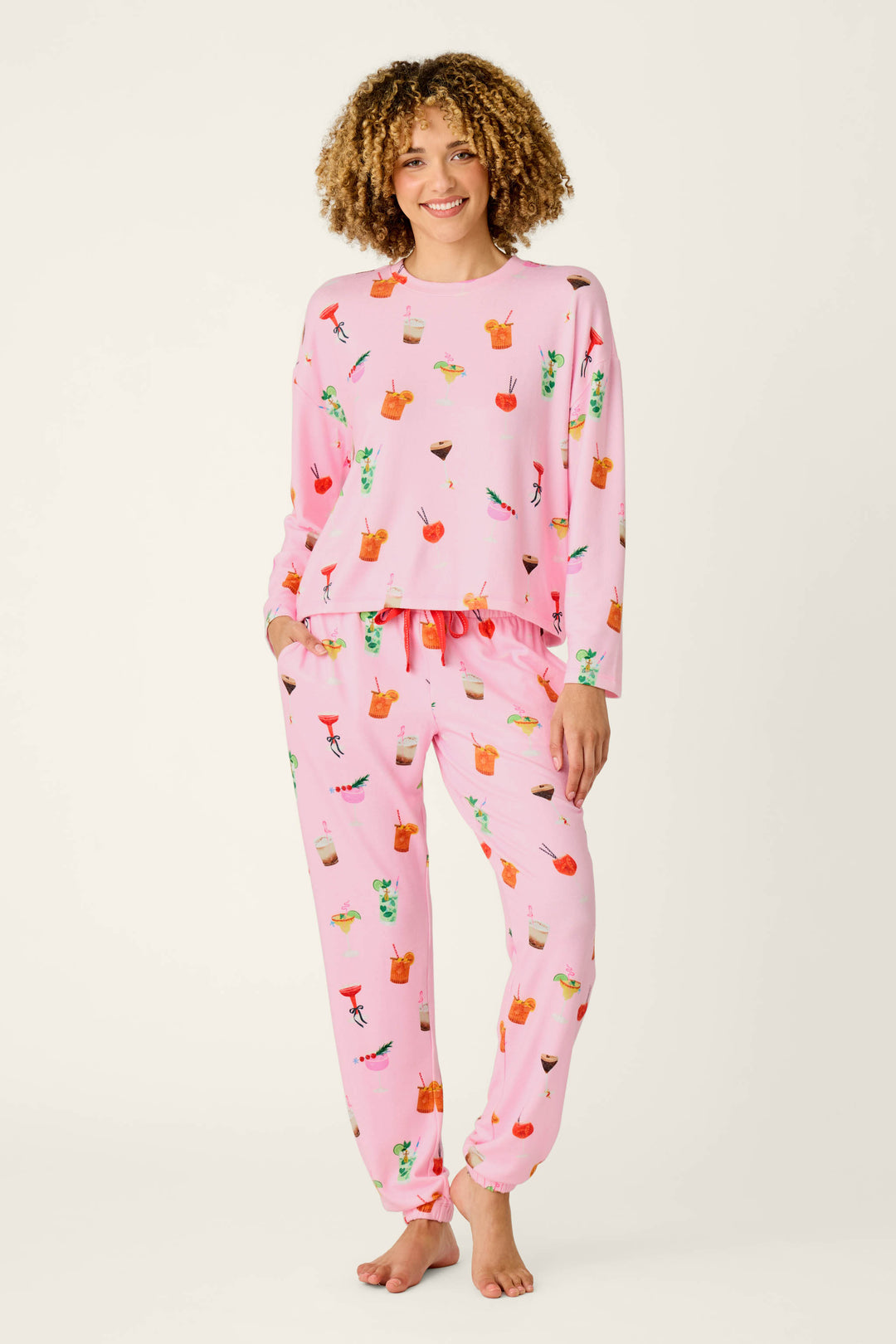A smiling person with curly hair models the P.J. Salvage First Chair, Last Call Cocktail Print Long Sleeve Top, featuring playful colorful fruit and drink designs, while standing barefoot against a plain light background.
