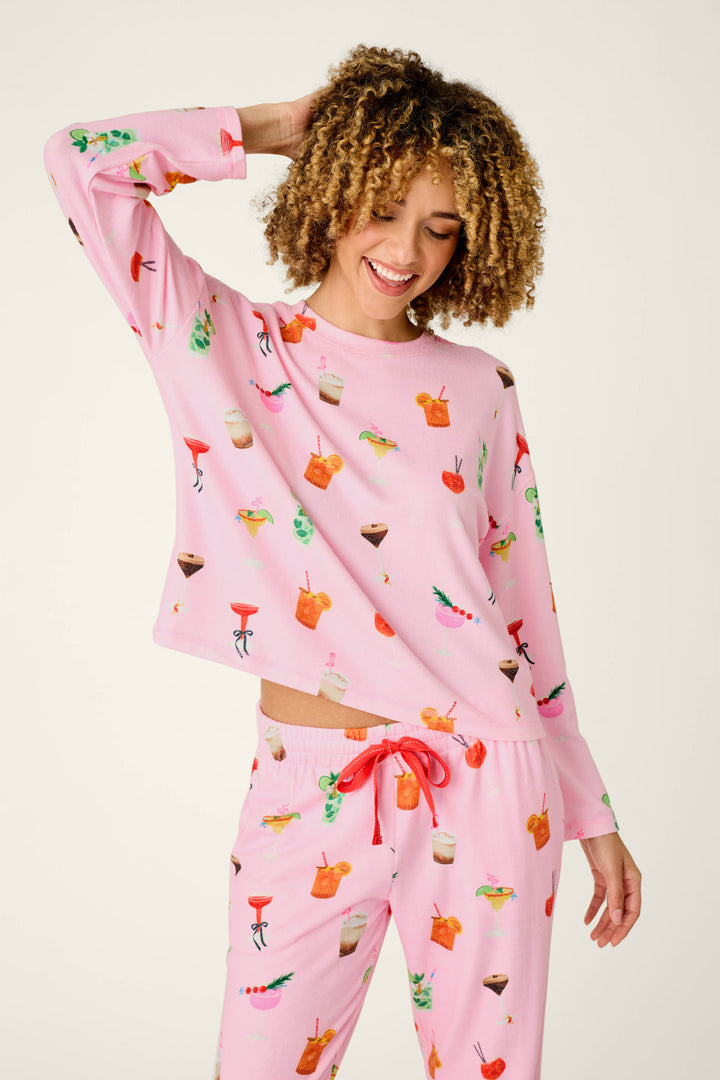 A person with curly hair smiles as they pose with one hand on their head, wearing the P.J. Salvage First Chair, Last Call Cocktail Print Long Sleeve Top—pink pajamas adorned with colorful cocktail and drink illustrations.
