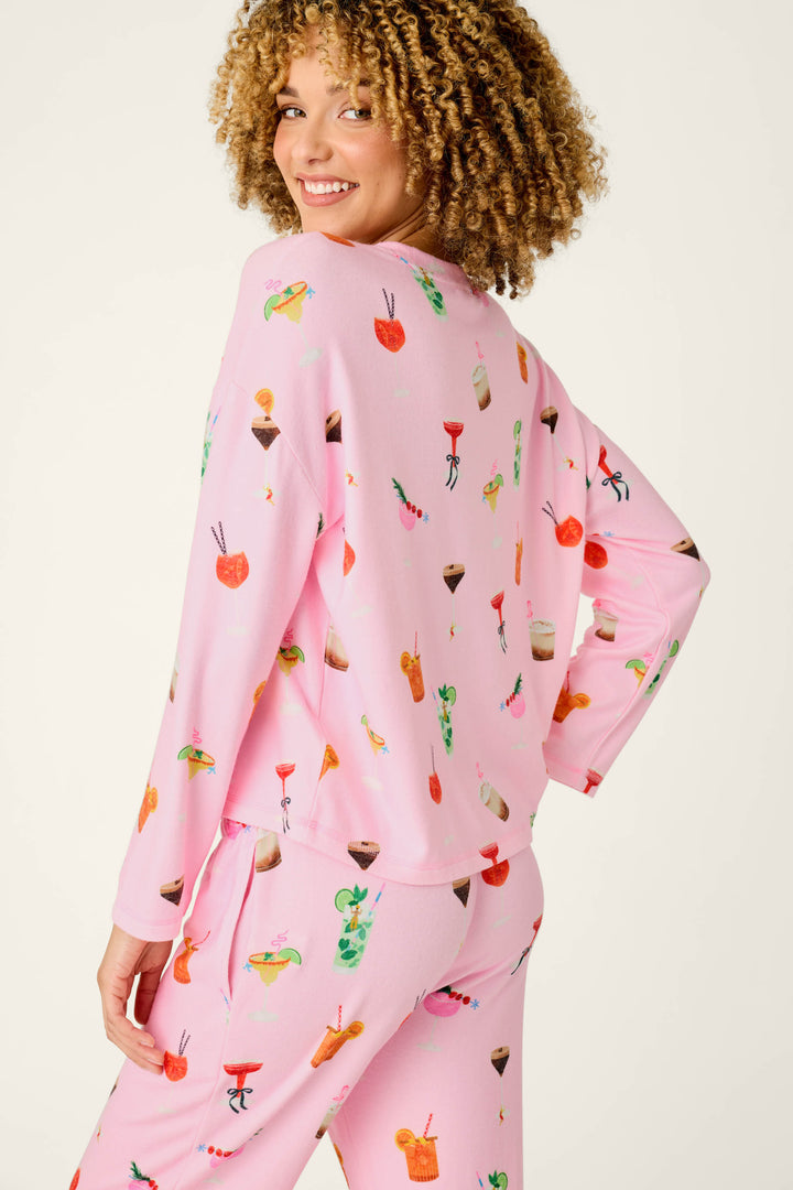 A person with curly hair smiles over their shoulder, wearing the P.J. Salvage First Chair, Last Call Cocktail Print Long Sleeve Top—pink pajamas featuring colorful drink and dessert illustrations on a light background.
