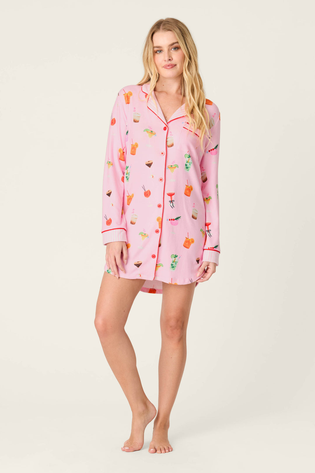 A woman stands barefoot against a plain light background, wearing the P.J. Salvage First Chair, Last Call Cocktail Print Long Sleeve Nightshirt featuring colorful drink and fruit patterns.