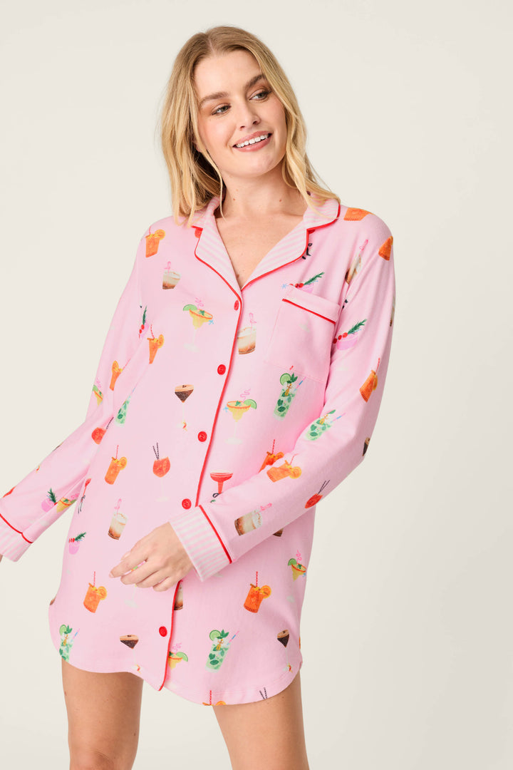 A woman smiles and looks to the side against a plain light background, wearing the P.J. Salvage First Chair, Last Call Cocktail Print Long Sleeve Nightshirt featuring a pink fabric with colorful cocktail motifs and red piping accents.