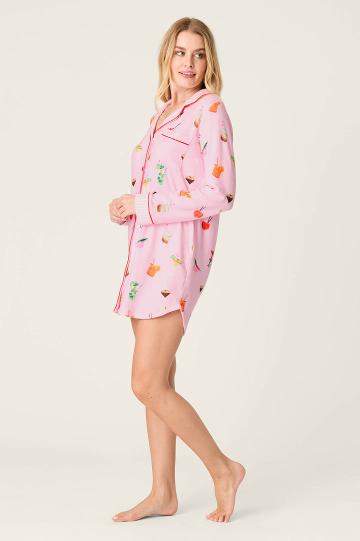 A woman, barefoot and smiling, stands sideways against a plain light background wearing the P.J. Salvage First Chair, Last Call Cocktail Print Long Sleeve Nightshirt— a pink pajama shirt adorned with colorful fruit and drink illustrations.