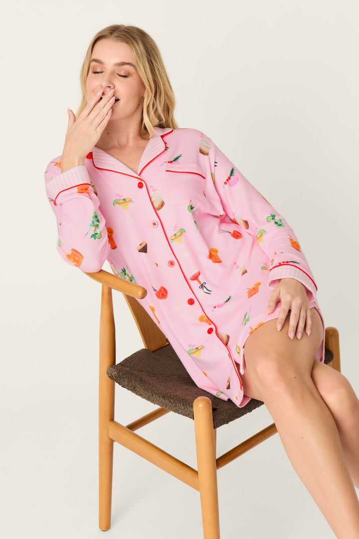 A woman wearing the P.J. Salvage First Chair, Last Call Cocktail Print Long Sleeve Nightshirt sits on a wooden chair, appearing to yawn with her eyes closed and hand over her mouth.