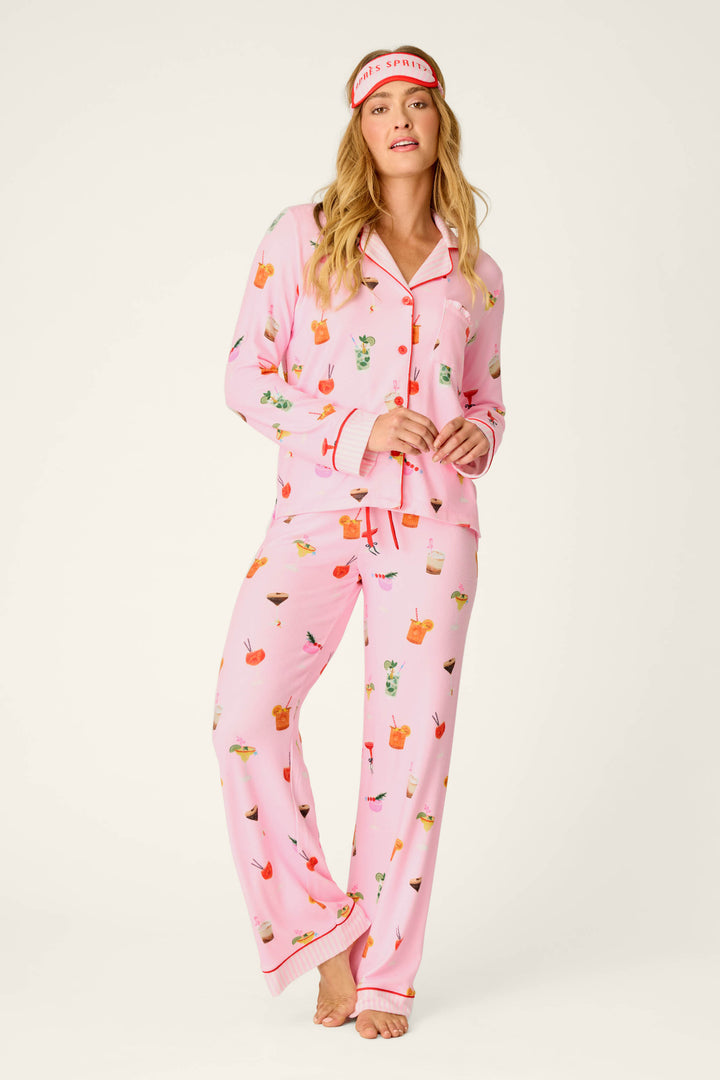 A woman stands barefoot and smiling in a P.J. Salvage First Chair, Last Call Cocktail Print 3-Piece PJ Set. She has a red-and-white sleep mask from the set pushed up on her forehead. The background is plain and light-colored.