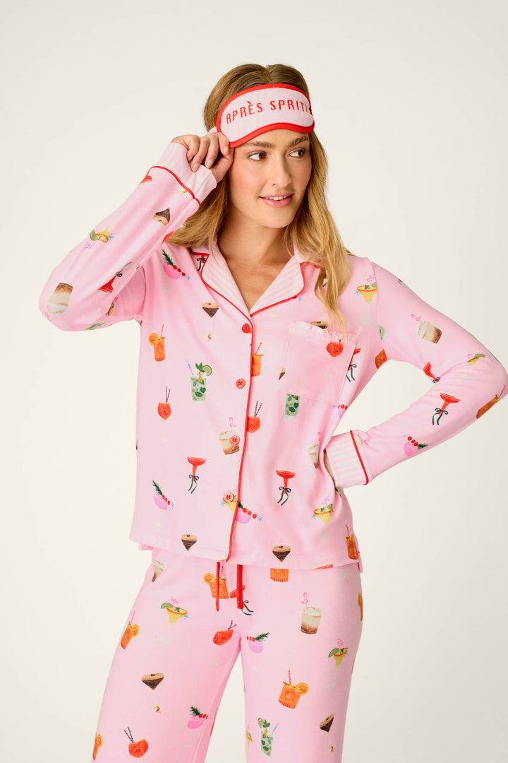 A woman models the P.J. Salvage First Chair, Last Call Cocktail Print 3-Piece PJ Set, featuring a pink pajama set with colorful cocktail illustrations and a matching pink sleep mask embroidered with “APRÈS SPRITZ” in red letters, standing against a simple light background.