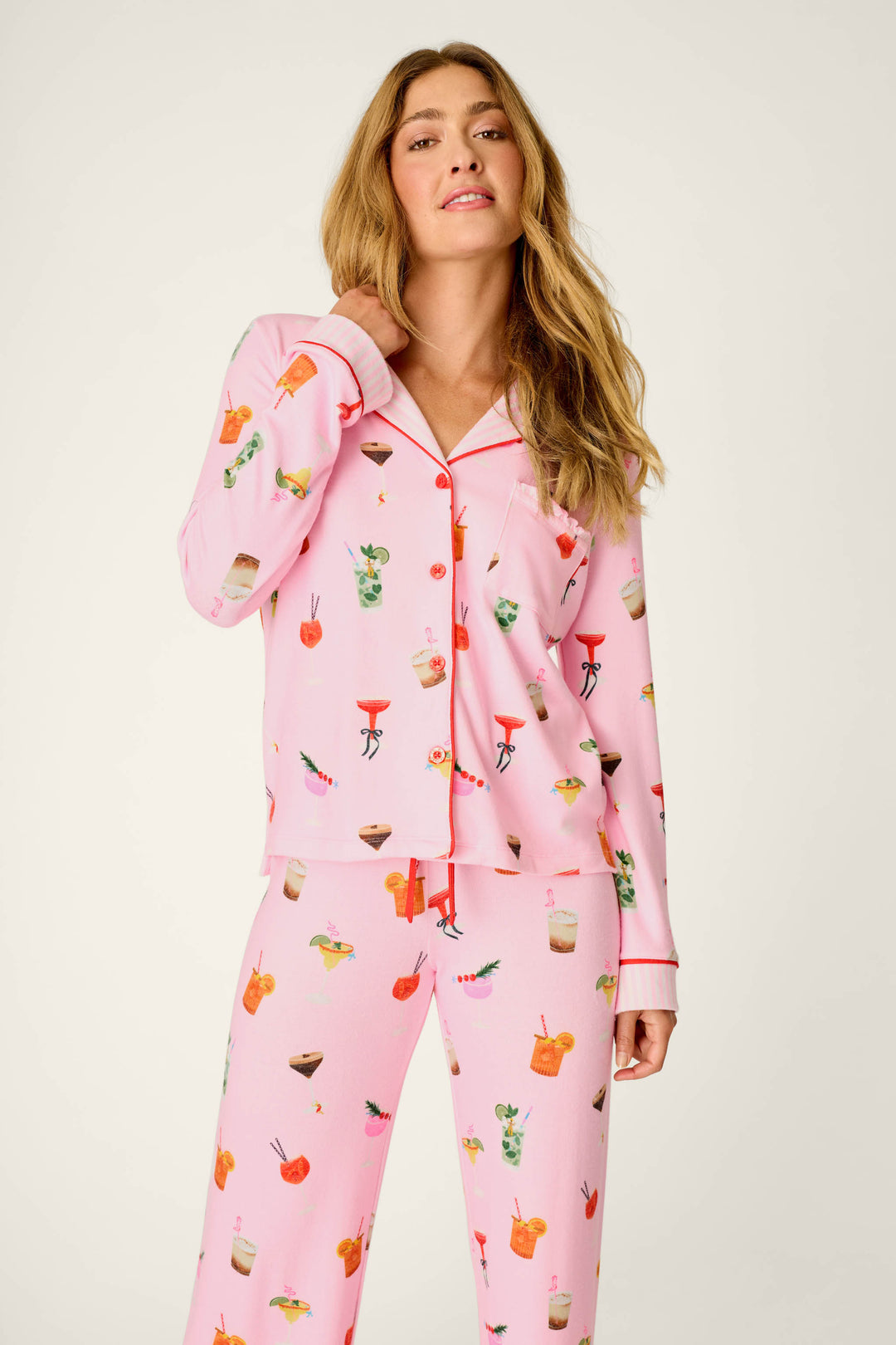 A woman with long, wavy blonde hair and a relaxed expression stands against a plain light background, wearing the P.J. Salvage First Chair, Last Call Cocktail Print 3-Piece PJ Set in light pink with colorful cocktail and fruit designs. One hand gently touches her collar.