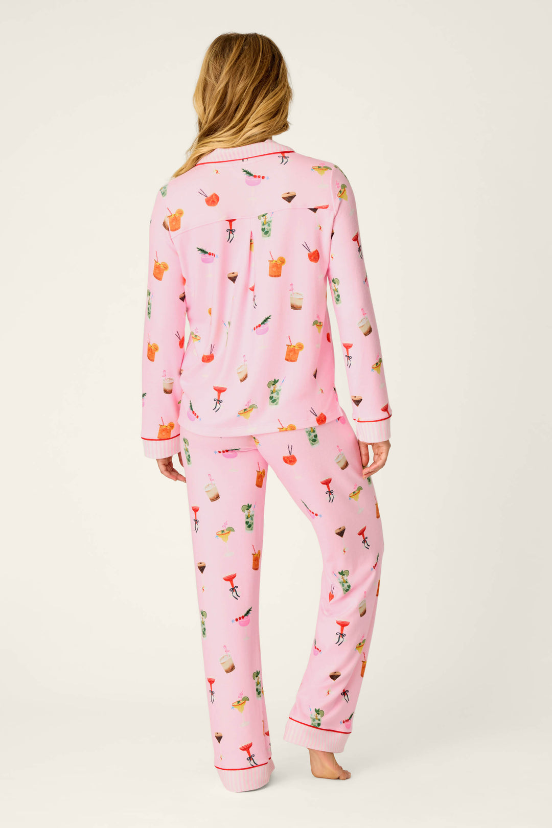 A person with long, light brown hair is seen from behind, standing barefoot against a plain background while wearing the P.J. Salvage First Chair, Last Call Cocktail Print 3-Piece PJ Set—pink pajamas adorned with colorful drink illustrations.