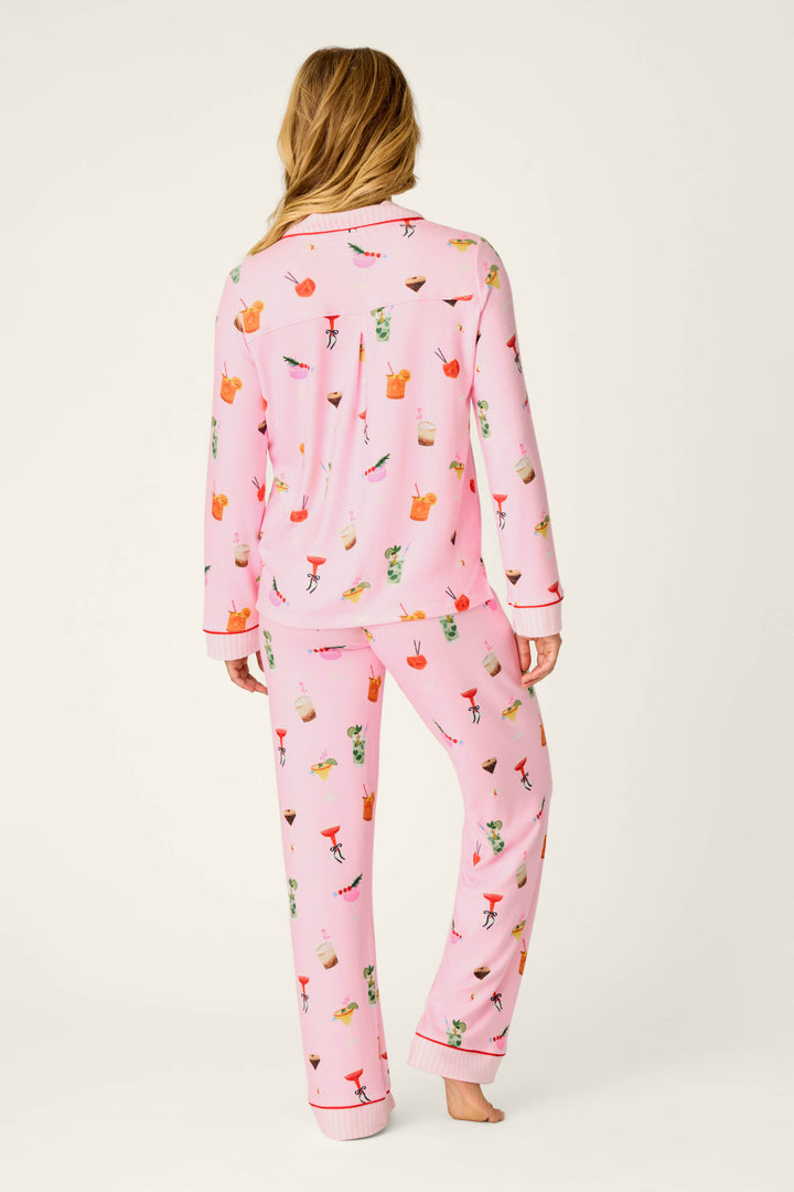 A person with long, light brown hair is seen from behind, standing barefoot against a plain background while wearing the P.J. Salvage First Chair, Last Call Cocktail Print 3-Piece PJ Set—pink pajamas adorned with colorful drink illustrations.