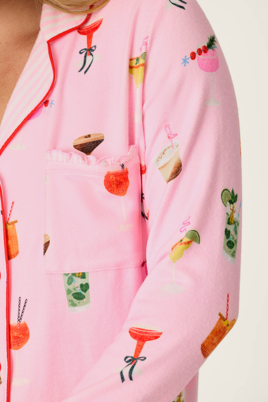 A close-up of a person wearing the P.J. Salvage First Chair, Last Call Cocktail Print 3-Piece PJ Set, featuring playful colorful drink illustrations including cocktails, smoothies, and fruit garnishes. The set includes pink pajamas with a chest pocket accented by a ruffled edge.