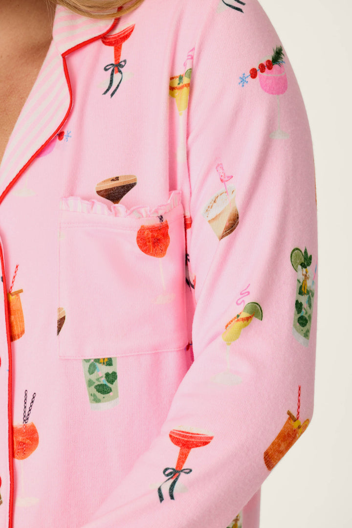 A close-up of a person wearing the P.J. Salvage First Chair, Last Call Cocktail Print 3-Piece PJ Set, featuring playful colorful drink illustrations including cocktails, smoothies, and fruit garnishes. The set includes pink pajamas with a chest pocket accented by a ruffled edge.