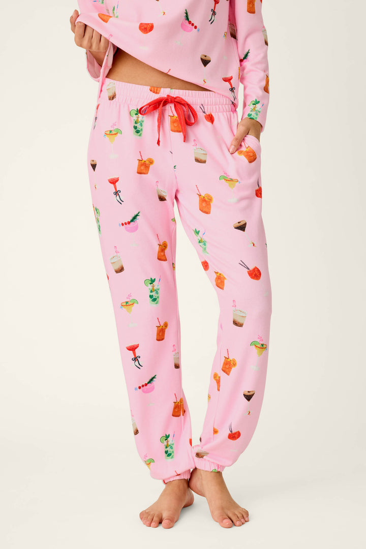 A person is wearing the P.J. Salvage First Chair, Last Call Cocktail Print Jogger Pajama Pants featuring colorful cocktail and drink illustrations in light pink, standing barefoot with only the lower body visible; a matching pajama top is partially shown.