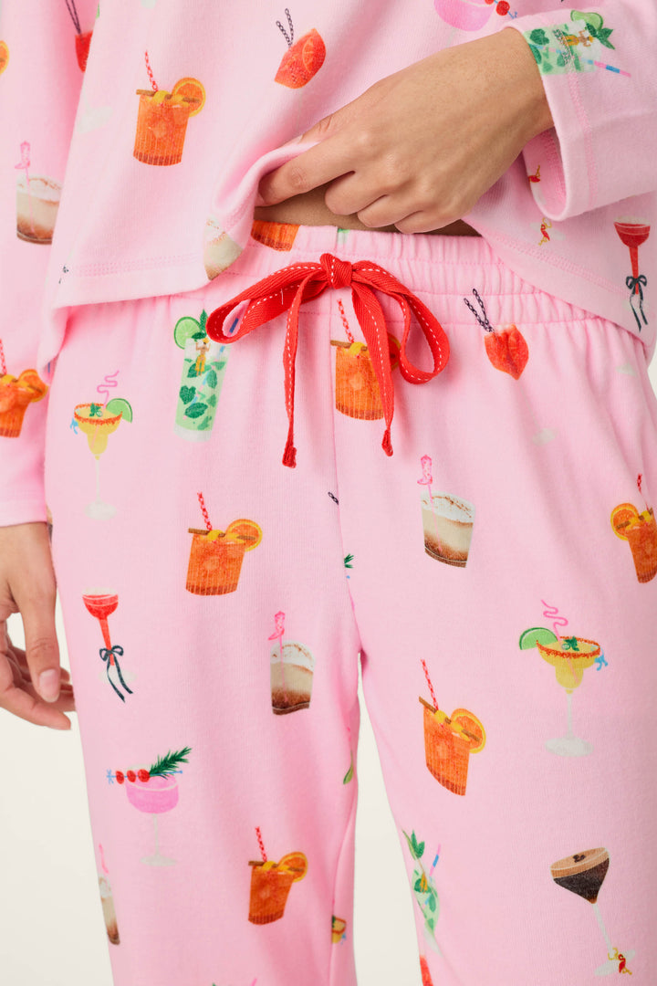 A person wearing P.J. Salvage’s First Chair, Last Call Cocktail Print Jogger Pajama Pants in pink—adorned with colorful cocktail illustrations and featuring a red drawstring—is shown from the torso to upper legs, with one hand resting near the waistband.