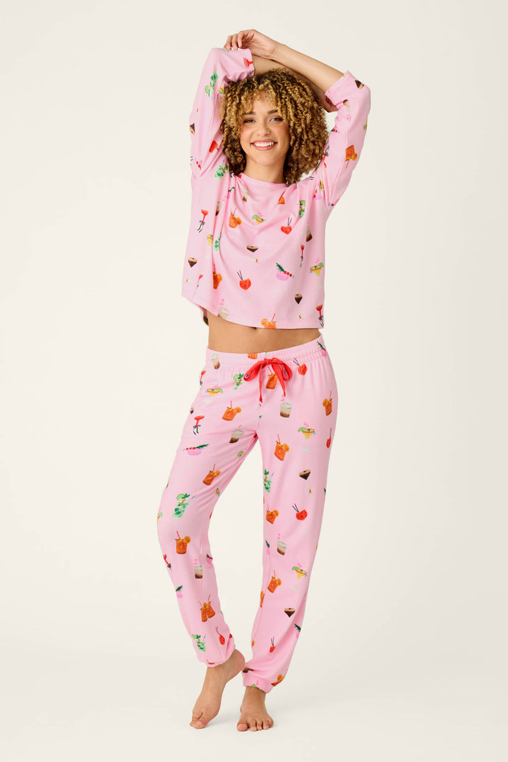 A smiling person with curly hair wears P.J. Salvage’s First Chair, Last Call Cocktail Print Jogger Pajama Pants in matching pink, featuring colorful cocktail-themed prints, standing barefoot with arms raised and one leg bent against a plain light background.