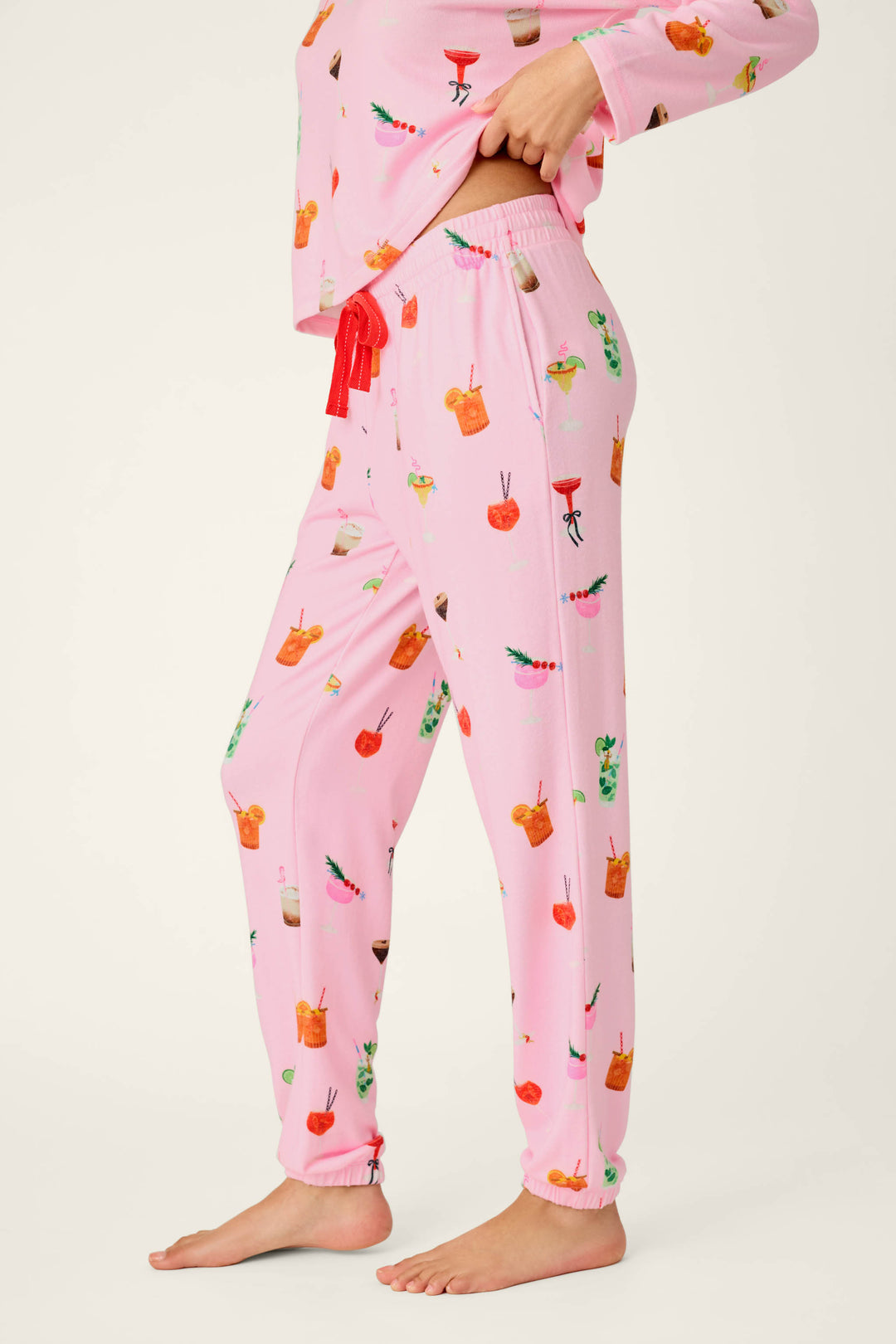 A person stands barefoot and sideways against a plain light background, wearing P.J. Salvage’s First Chair, Last Call Cocktail Print Jogger Pajama Pants featuring colorful cocktail and fruit designs, with the matching top partially visible.