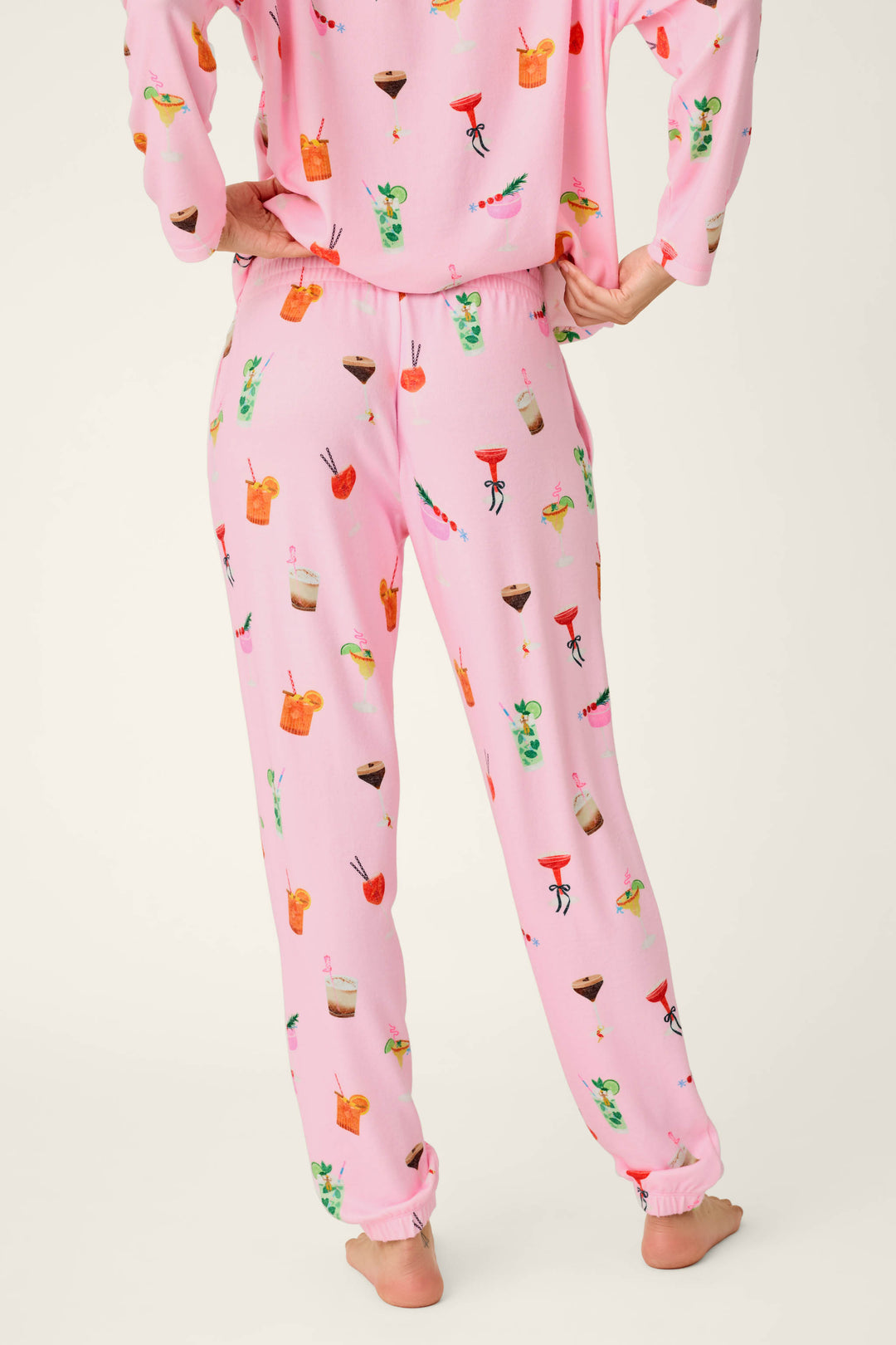 A person stands barefoot, shown from the back, wearing P.J. Salvage’s First Chair, Last Call Cocktail Print Jogger Pajama Pants featuring colorful cocktail and drink illustrations. The matching pajama top is partially visible against a plain off-white background.