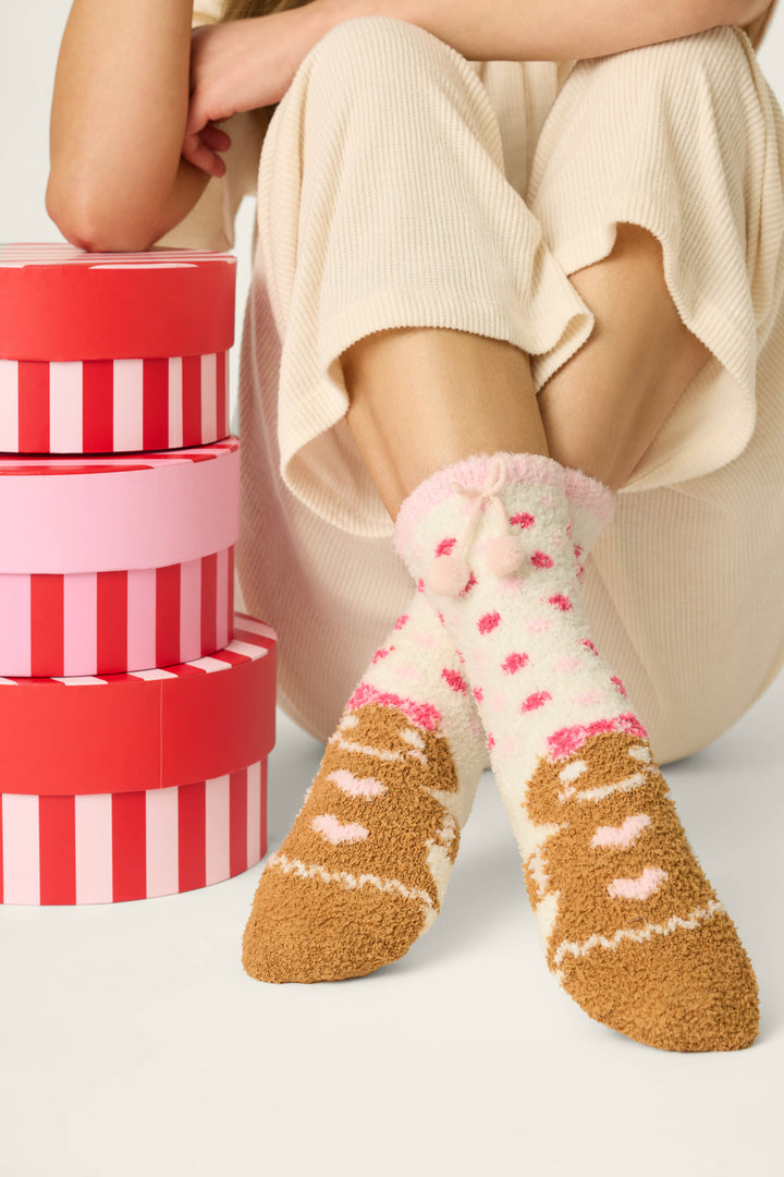 Wearing P.J. Salvage's Gingerbread Girl & Polka Dots Socks, a person with cozy pom-pom accents sits next to a stack of red and pink striped gift boxes.