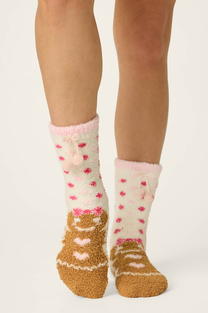 A person wearing P.J. Salvage's Gingerbread Girl & Polka Dots Socks—one soft and warm sock features white with pink polka dots and playful pom-poms, while the other showcases a brown design adorned with pink and white heart patterns.