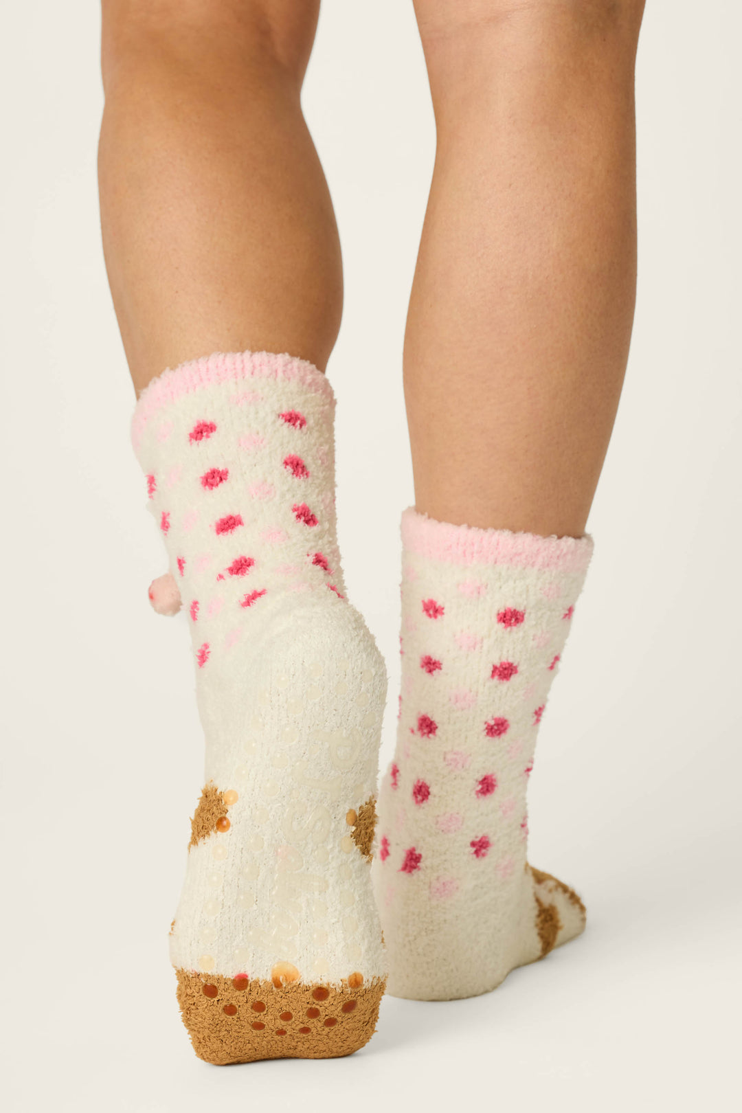 A person is wearing P.J. Salvage Gingerbread Girl & Polka Dots Socks, featuring fuzzy white fabric with pink polka dots and non-slip grips on the bottom, standing on a white background.
