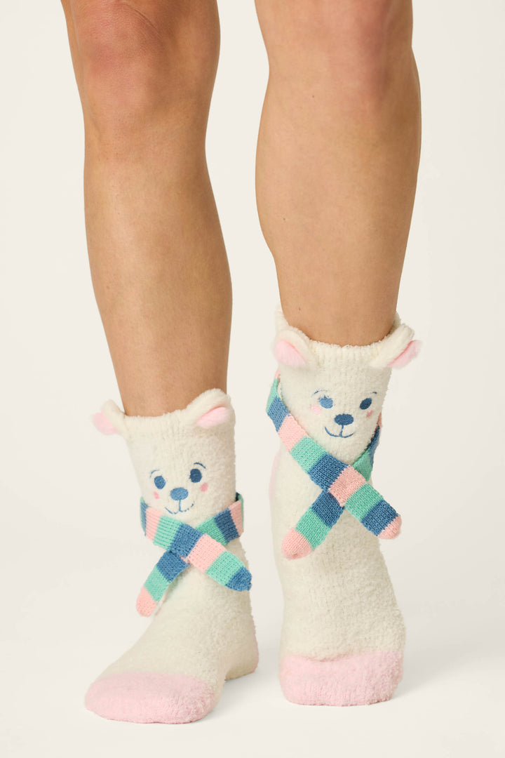 Two legs wearing P.J. Salvage Cozy Polar Bear Socks, fuzzy white socks designed to resemble bears with pink ears, blue eyes, multicolored scarves, and pink toes, standing out against a plain background.