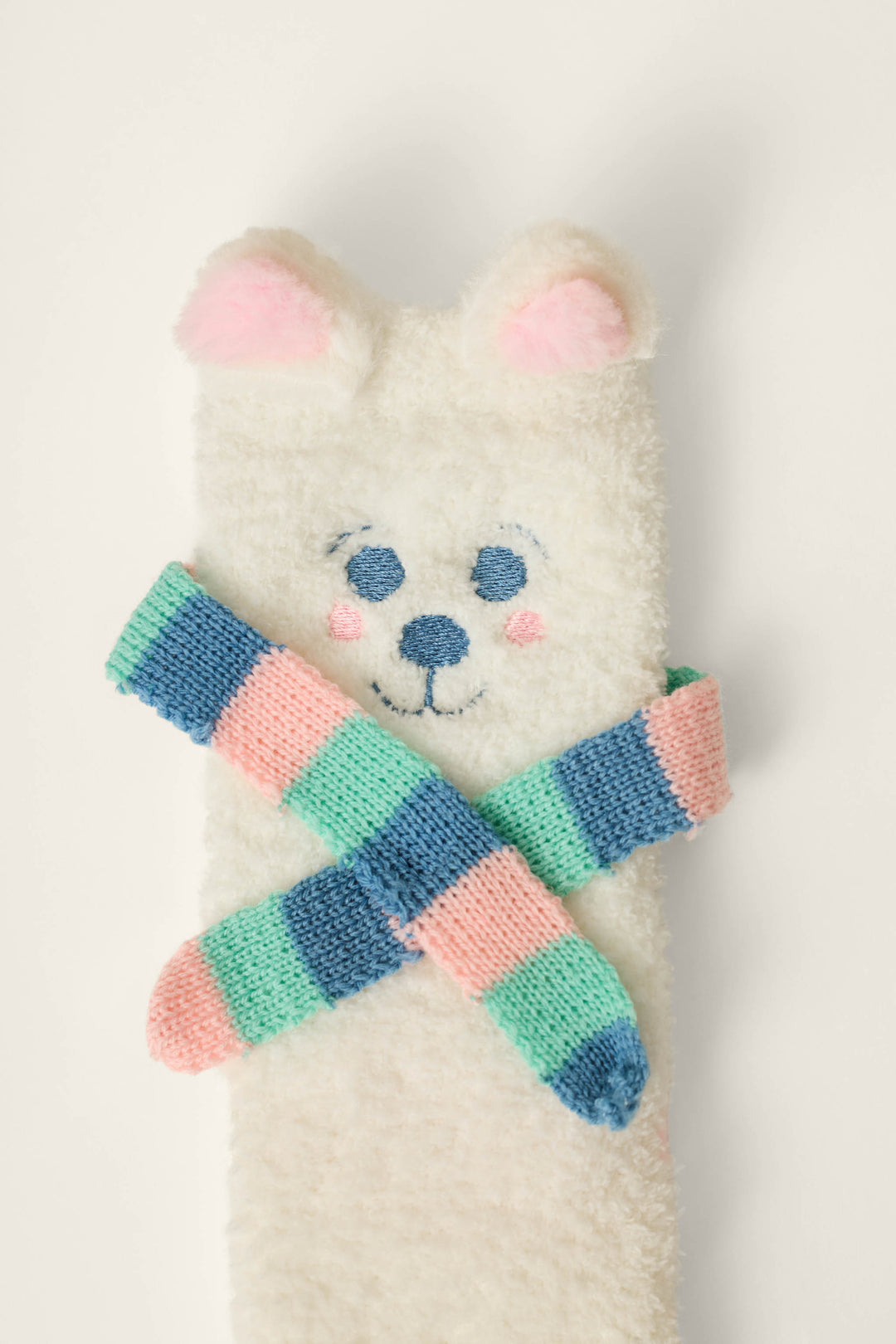 On a plain background, the P.J. Salvage Cozy Polar Bear Socks—featuring a fuzzy white design with a bear face, pink ears, and blushing cheeks—are paired with striped blue, green, and pink socks crossed in front of them.