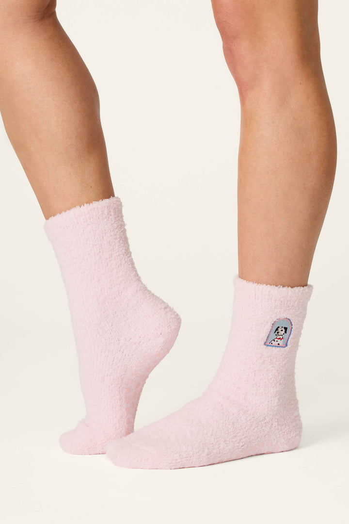 Two legs wearing P.J. Salvage's "Puppy Portrait Socks" in fuzzy, light pink fabric that reach mid-calf; one sock displays a small cartoon dog face patch on the outer side. The background is plain and light-colored.