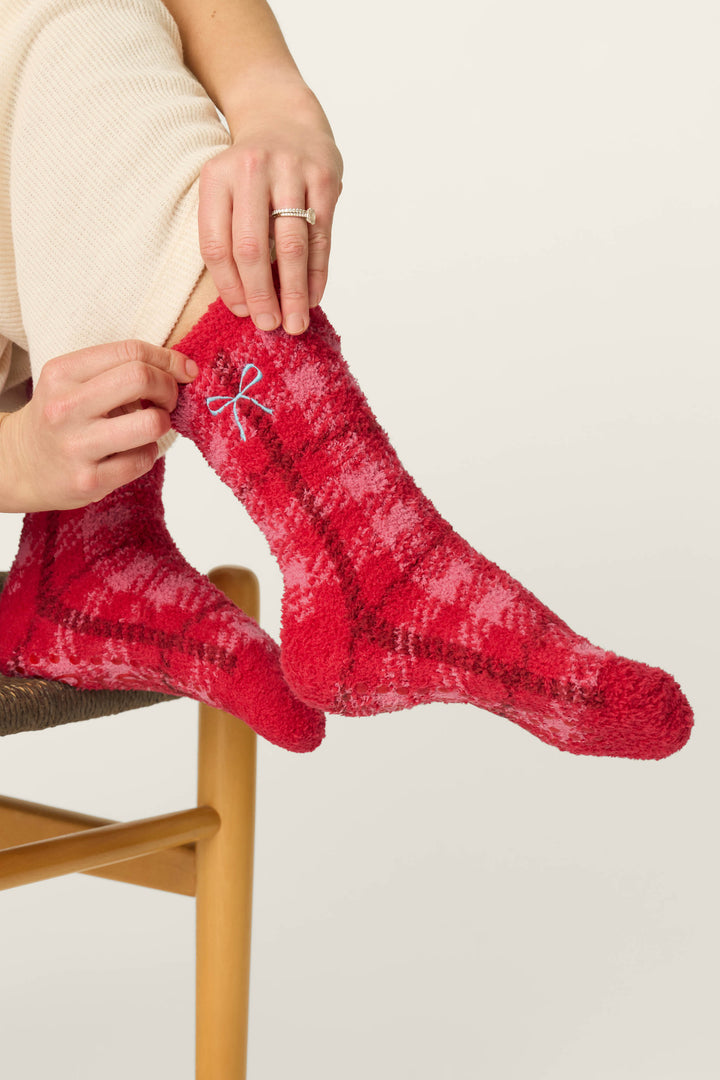 A person sitting on a chair pulls up a pair of P.J. Salvage Red Plaid with Embroidered Bow Socks featuring a small blue bow. They are wearing cream-colored pants and a silver ring on their left hand.
