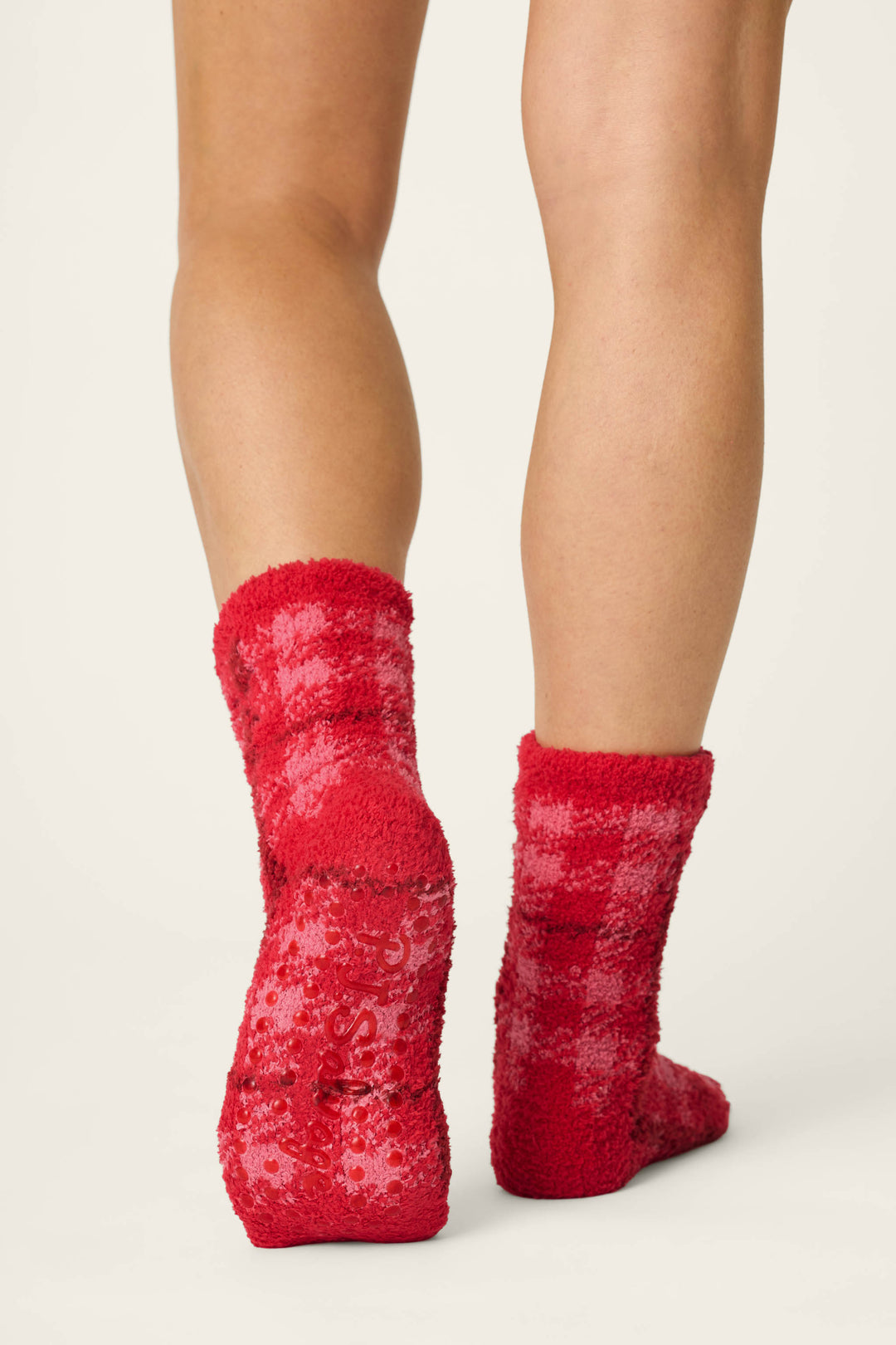 A person stands on a light surface wearing P.J. Salvage Red Plaid with Embroidered Bow Socks, featuring grip dots on the soles. The image is centered on the back of their lower legs and feet.
