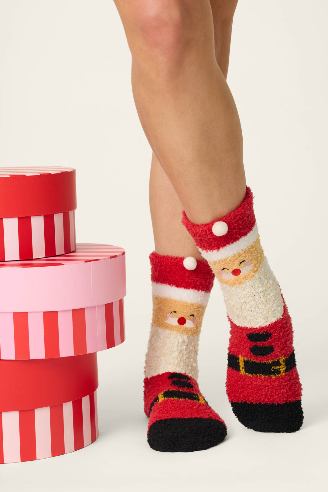 Wearing P.J. Salvage’s Jolly Santa Claus Socks—festive fuzzy socks in red, white, and black with cheerful Santa faces—a person stands beside a stack of round gift boxes with pink and red stripes on a light background.