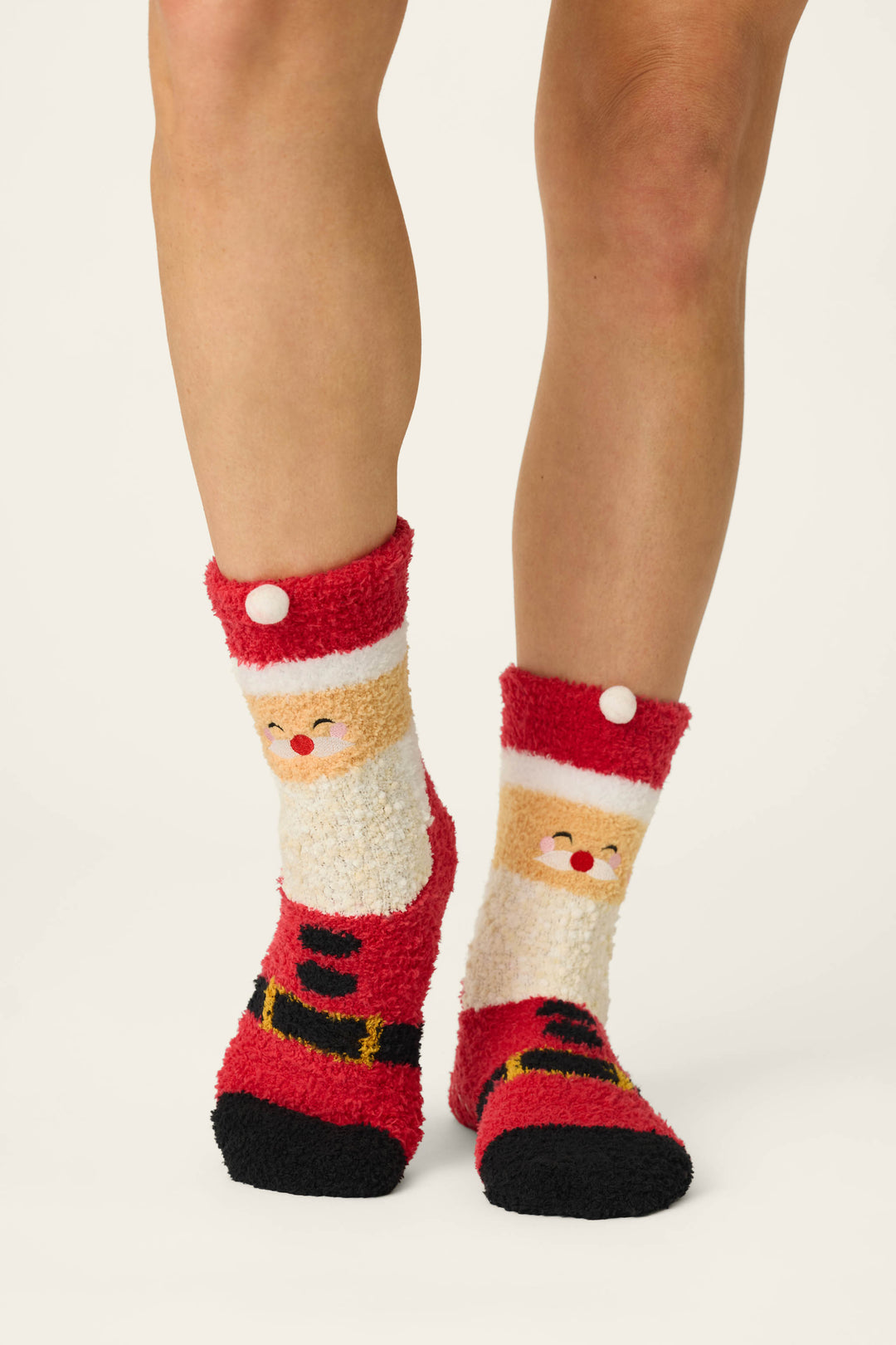 A person wears the P.J. Salvage Jolly Santa Claus Socks—fuzzy, red socks adorned with Santa faces and belts. Only their lower legs and feet are visible against a plain, light background.