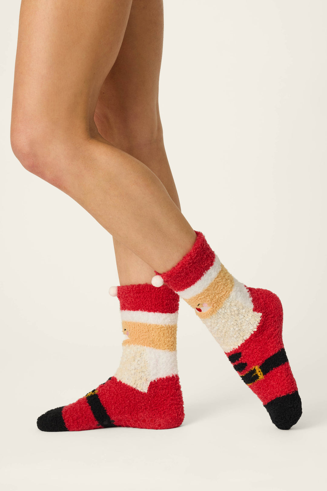 A person is wearing the P.J. Salvage Jolly Santa Claus Socks—cozy red, white, and beige socks featuring a fuzzy texture and Santa belt design—while sitting with crossed legs against a plain white background.