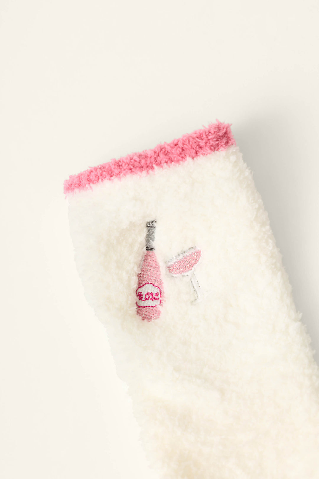 The Sip, Sip, Sparkle Embroidered Socks by P.J. Salvage are crafted from a fluffy white fabric with a pink fuzzy edge and feature embroidered designs of a pink champagne bottle and a glass filled with pink liquid.