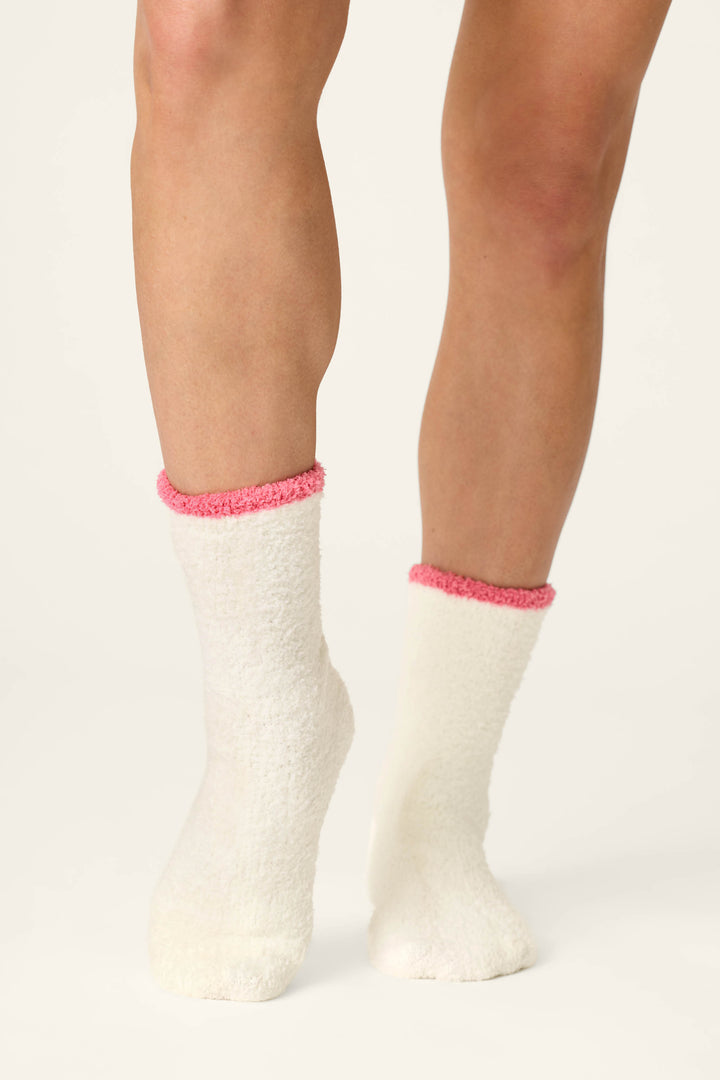 A person wears the P.J. Salvage Sip, Sip, Sparkle Embroidered Socks—white and fuzzy with pink trim—standing on a light background, with only their lower legs and feet visible.