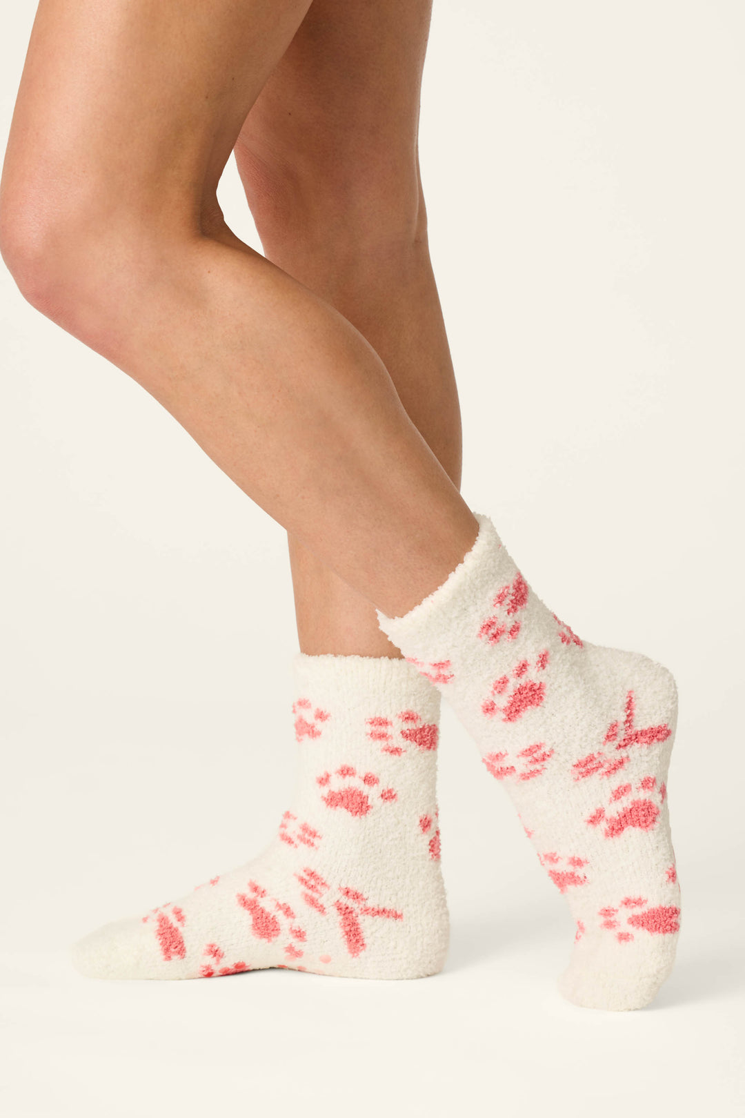 A person wearing comfy white Pink Paw Print Socks by P.J. Salvage stands with one foot forward, displaying both the side and back of the socks—featuring pink paw print designs—against a light background.