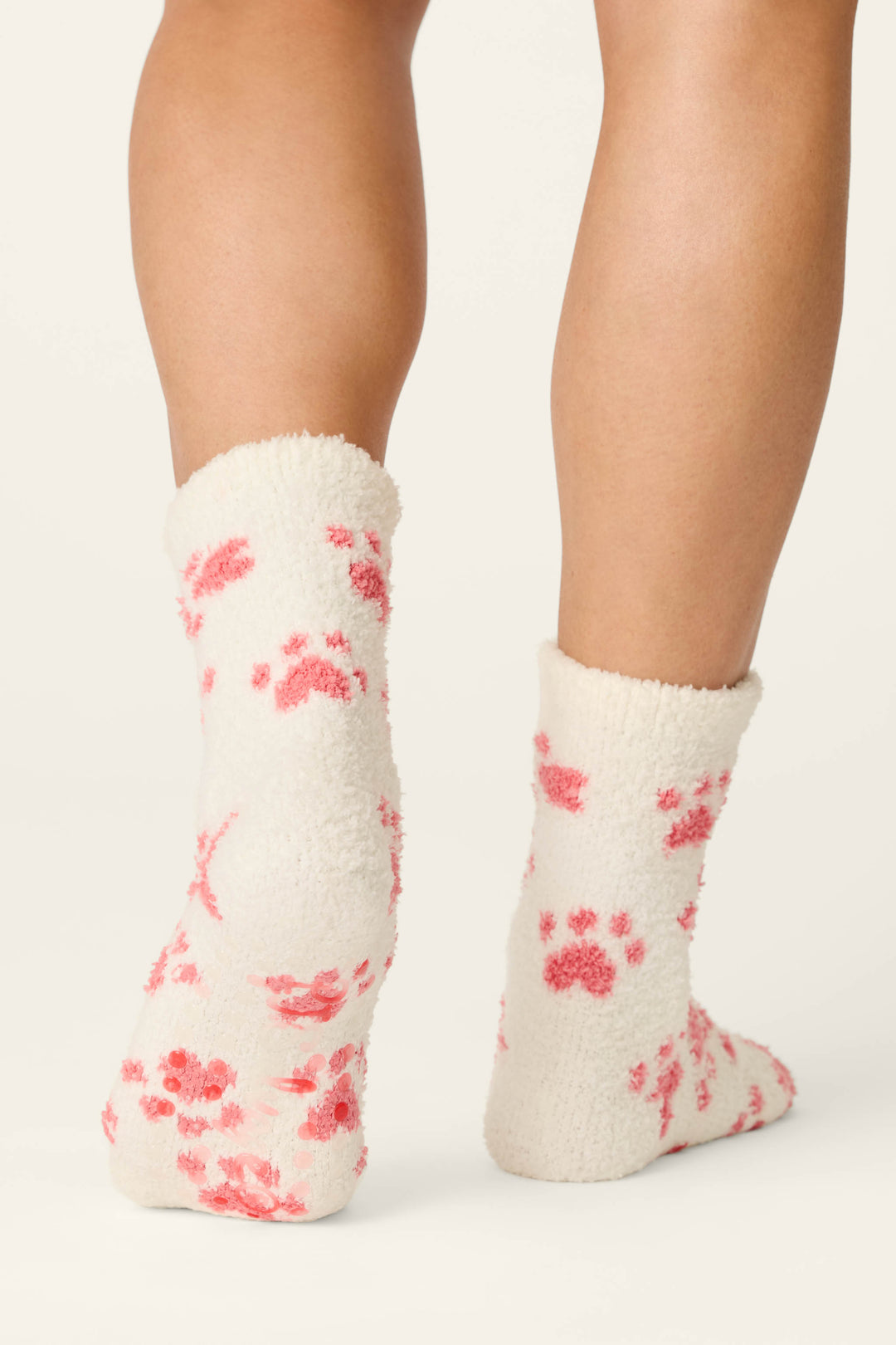 A person wears P.J. Salvage Pink Paw Print Socks, featuring a fuzzy white design with pink paw prints. The photo displays the back of their lower legs and feet against a plain background.