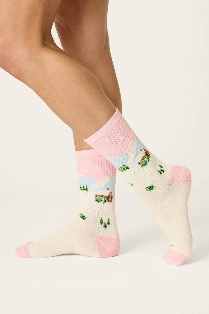 A person wearing P.J. Salvage Cozy Winter Cabin Crew Socks, which are cream-colored with pink heels, toes, and cuffs. The socks display a charming winter scene of houses, snow, mountains, and trees as the person’s legs are crossed at the ankle against a plain background.