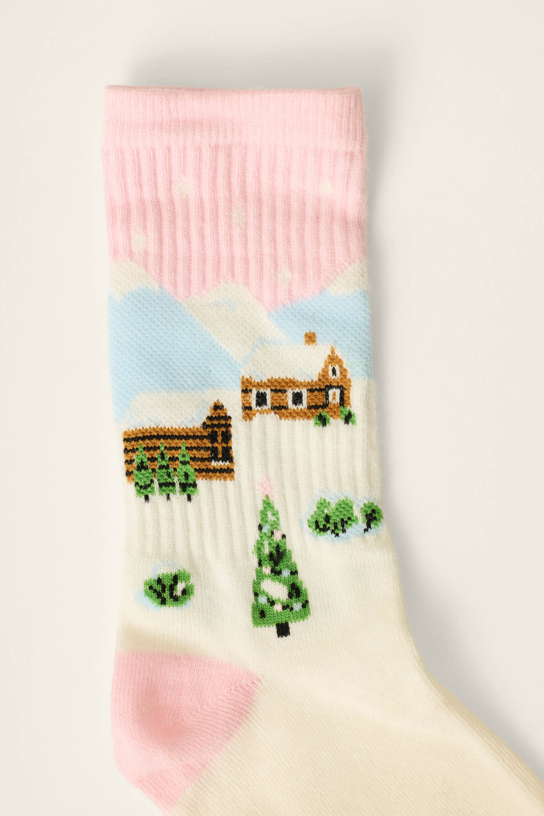 A single crew sock with a winter village pattern, featuring a pink ribbed cuff, a cream foot, and a printed design of a cabin, trees, and mountains under a light blue sky.