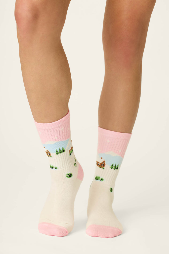 A person wearing P.J. Salvage Cozy Winter Cabin Crew Socks—cream-colored socks featuring pink toes, heels, and cuffs, adorned with charming houses, trees, and snow against a simple light background.