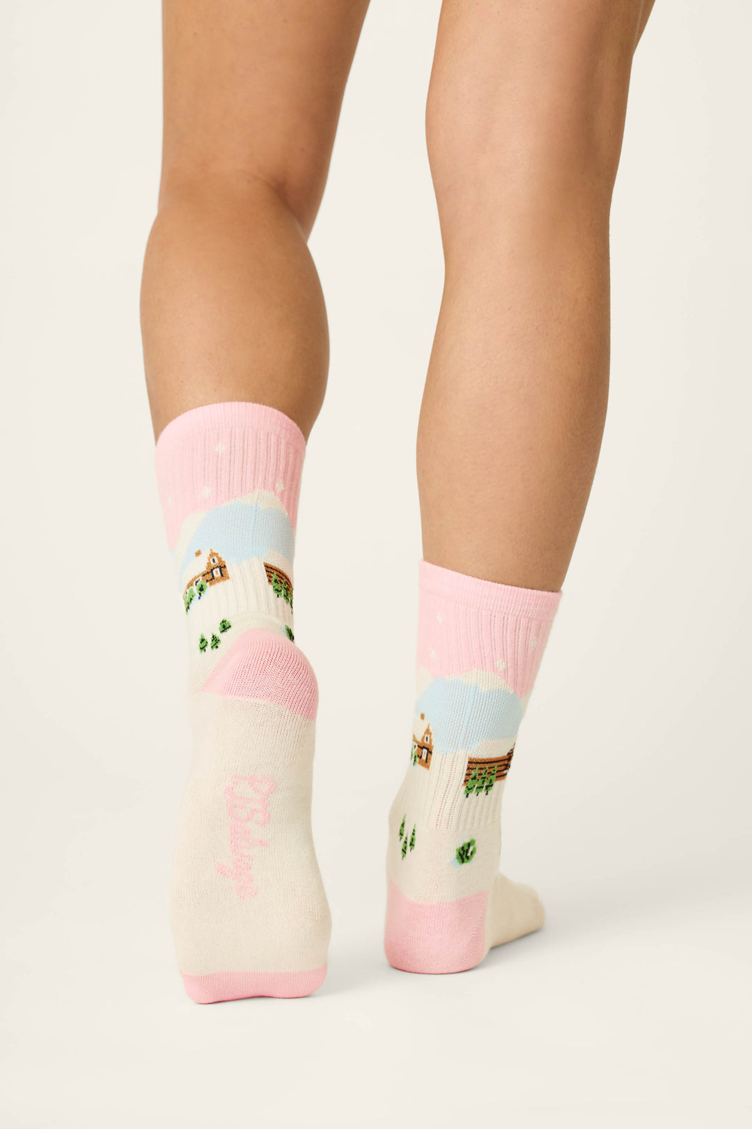 A person stands with their feet close together on a light floor, seen from behind, wearing P.J. Salvage Cozy Winter Cabin Crew Socks featuring a pink and white winter cabin and snow scene design.