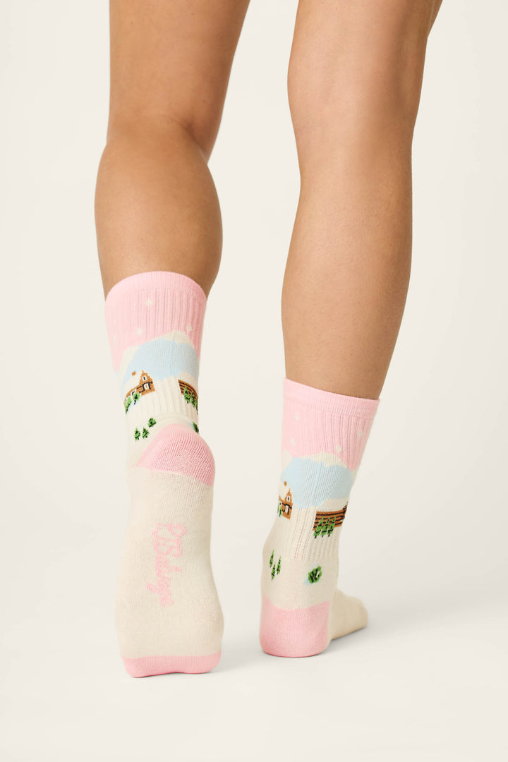 A person stands with their feet close together on a light floor, seen from behind, wearing P.J. Salvage Cozy Winter Cabin Crew Socks featuring a pink and white winter cabin and snow scene design.