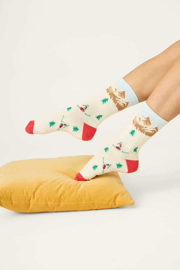 Two feet wearing P.J. Salvage Snowy Mountainscape Crew Socks, featuring cream-colored fabric with Christmas tree and skiing person patterns, red toes and heels, and mountain designs at the top, resting above a yellow pillow on a light background.